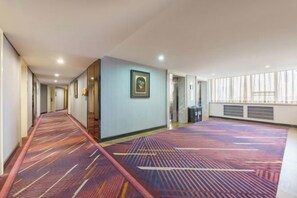 A hotel corridor with a patterned carpet, a framed picture on the wall, and a large window on the right.