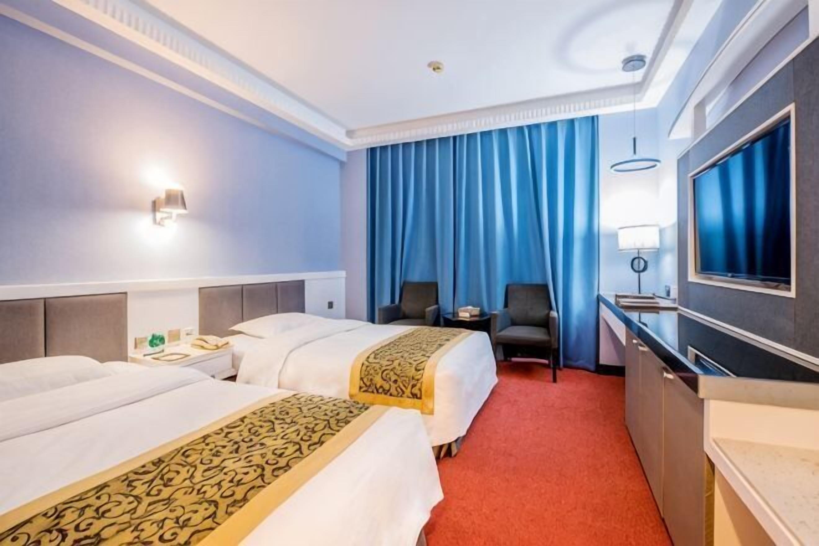 A hotel room with two beds, a desk, a chair, a TV, and a large window with curtains.