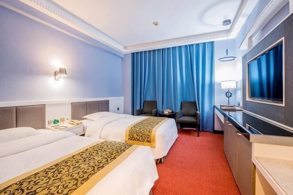 A hotel room with two beds, a desk, a chair, a TV, and a large window with curtains.