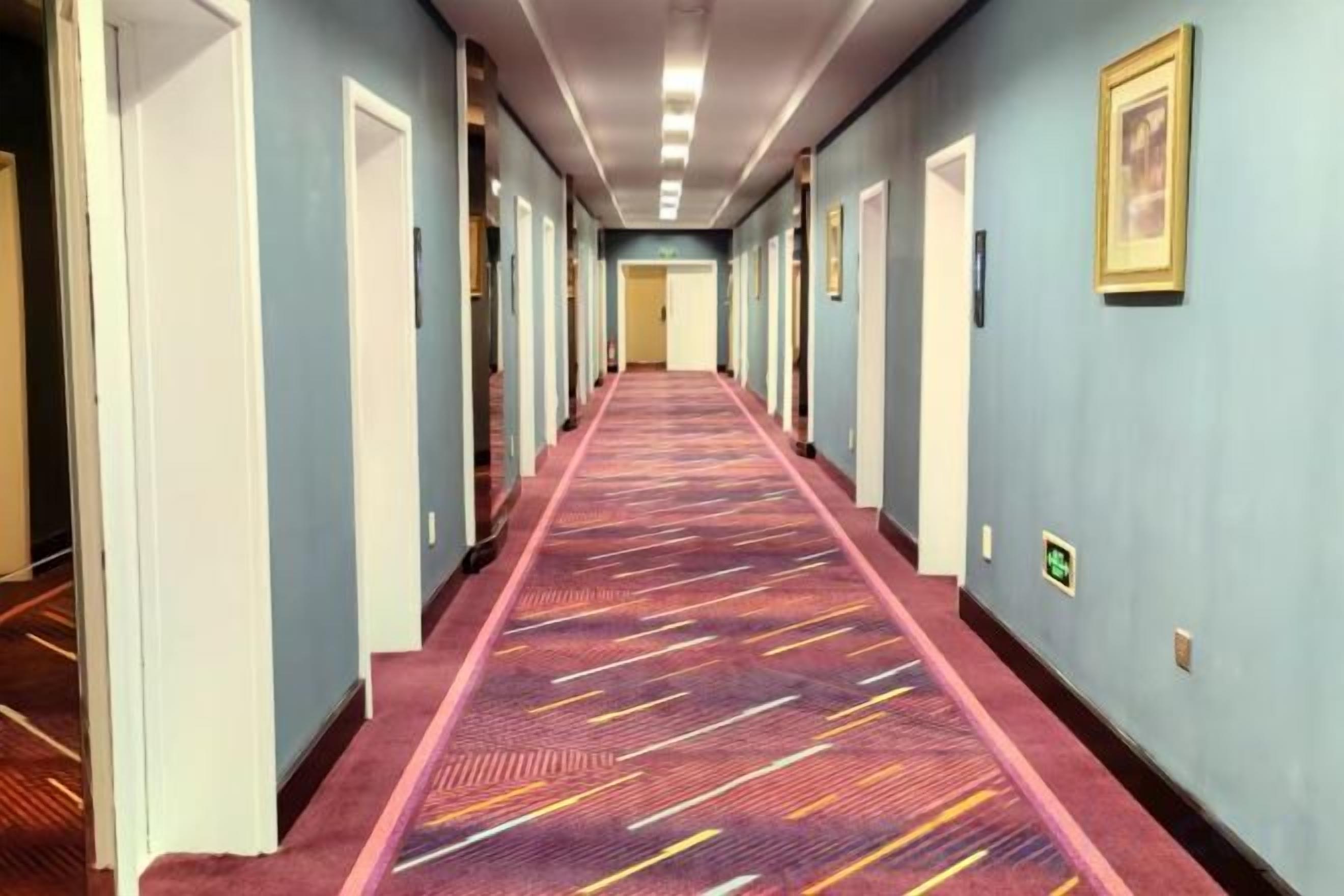 Ein Hotelgang mit einem gemusterten Teppich, Türen an beiden Seiten und gerahmten Bildern an den Wänden.