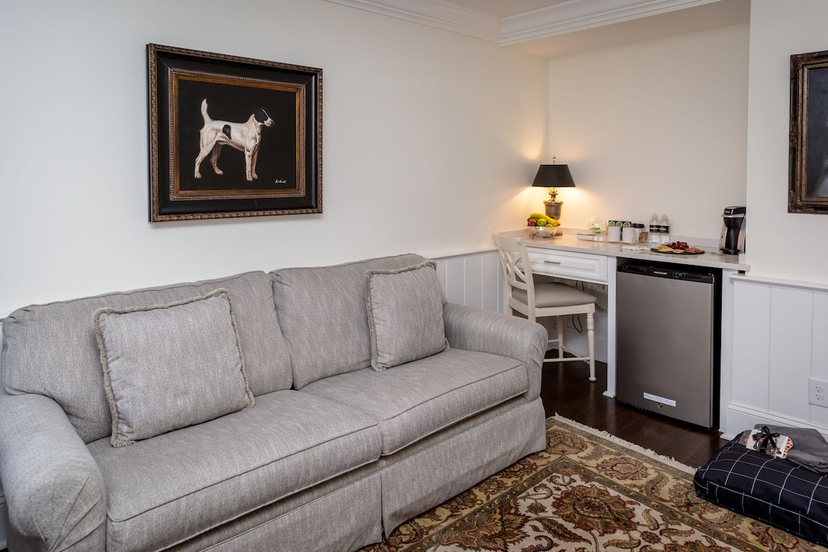 A living room with a grey sofa, a dining table, a lamp, and a kitchen area with a dishwasher.