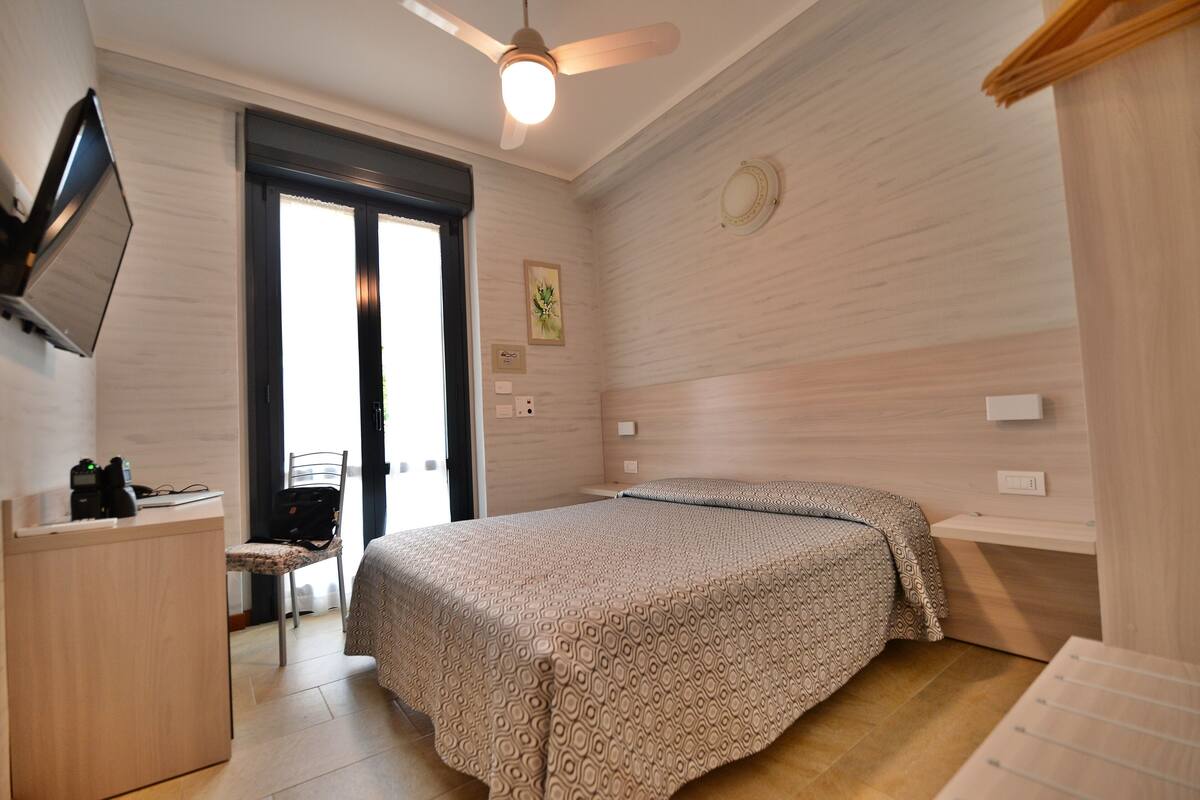 Een slaapkamer met een bed, een bureau, een stoel, een plafondventilator en een deur.