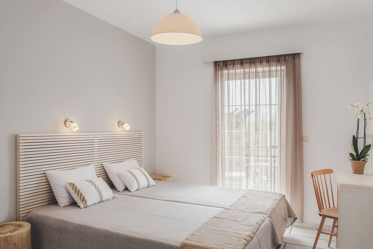 A modern bedroom with a large bed, bedside lamps, a wooden headboard, a window with sheer curtains, and a small desk with a chair.