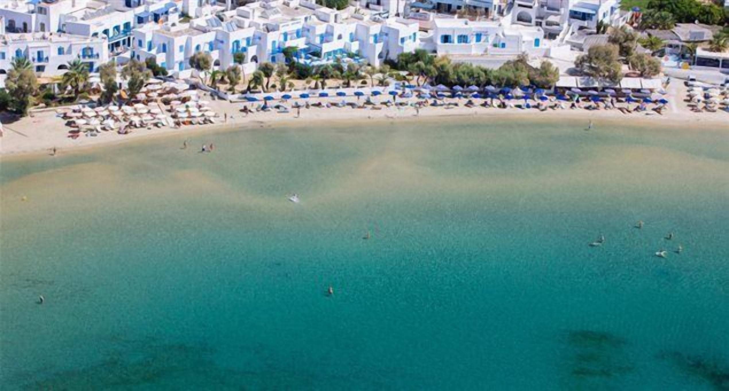 An aerial view of a coastal area with white buildings, a sandy beach, and umbrellas.
