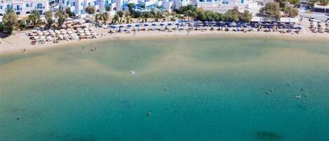 An aerial view of a coastal area with white buildings, a sandy beach, and umbrellas.
