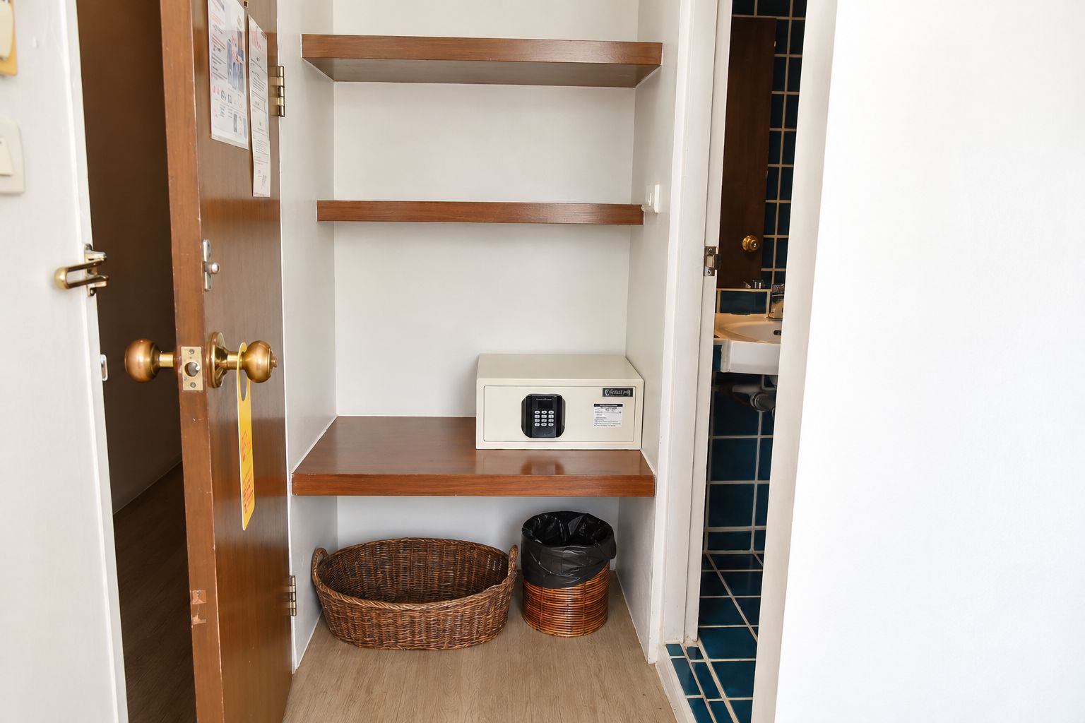 Un pasillo estrecho con una puerta a la izquierda, una pared blanca con un tablón de anuncios y una pared con azulejos azules a la derecha. Hay dos estantes de madera, uno de los cuales tiene un microondas.