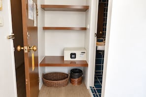 A narrow hallway with a door on the left, a white wall with a bulletin board, and a blue tiled wall on the right. There are two wooden shelves with a microwave on the lower shelf. - Villa Cha Cha Banglumphu (Bangkok)