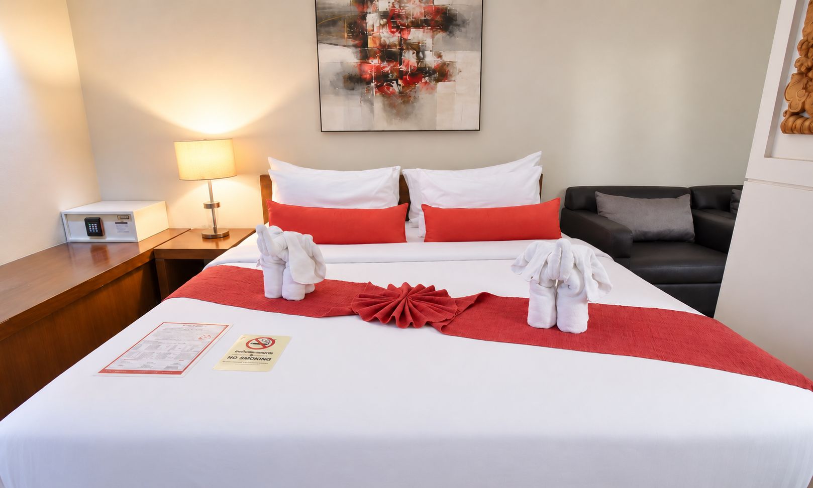 Une chambre d’hôtel avec un lit bien fait, un plaid rouge, des serviettes, un panneau « interdiction de fumer », une lampe, un tableau et un canapé.