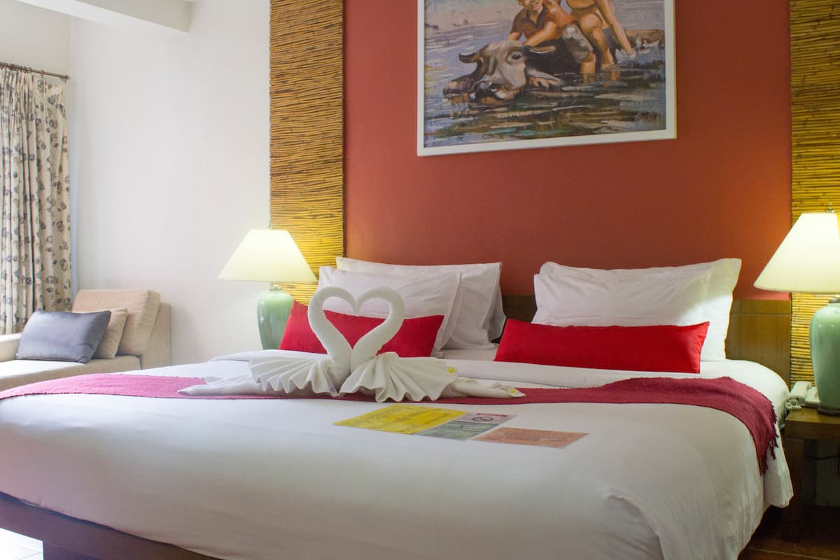 Een opgemaakt bed met wit en rood beddengoed, twee bedlampjes, een schilderij aan de muur en een bloemen gordijn.