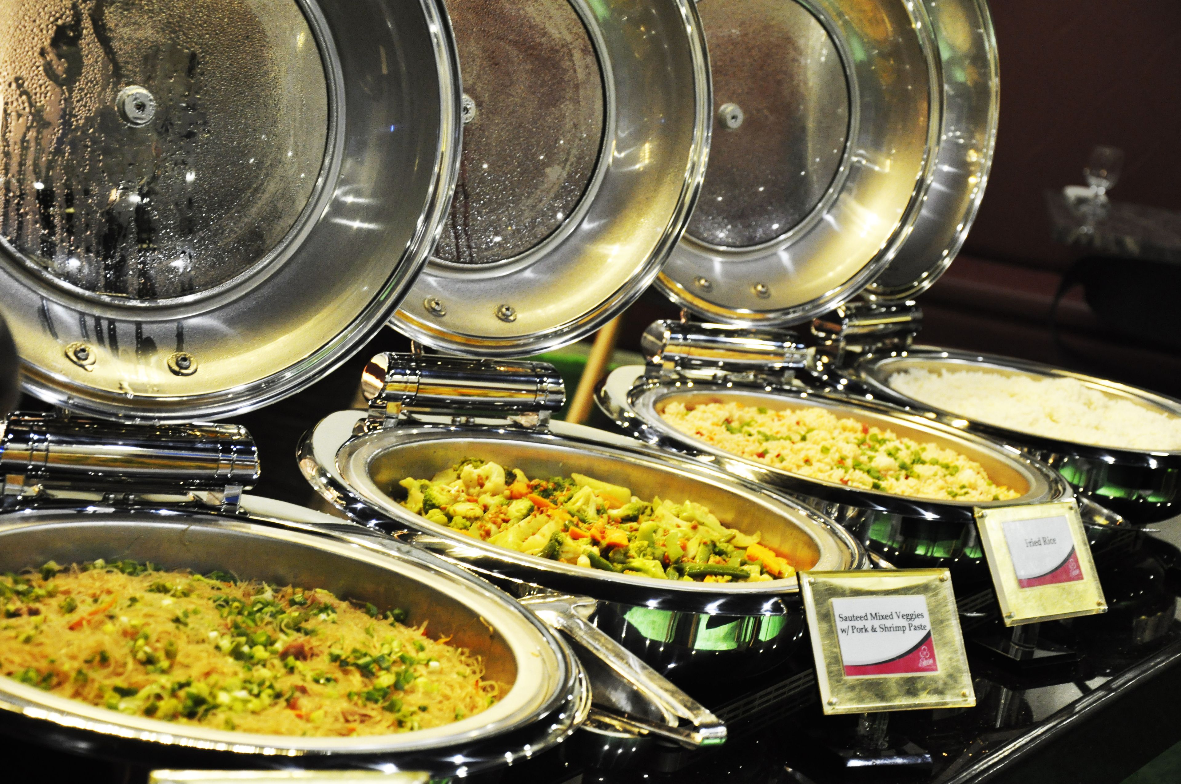 Een buffet met diverse gerechten, waaronder een bord met roerbakgroenten, varkensvlees en garnalenpasta.