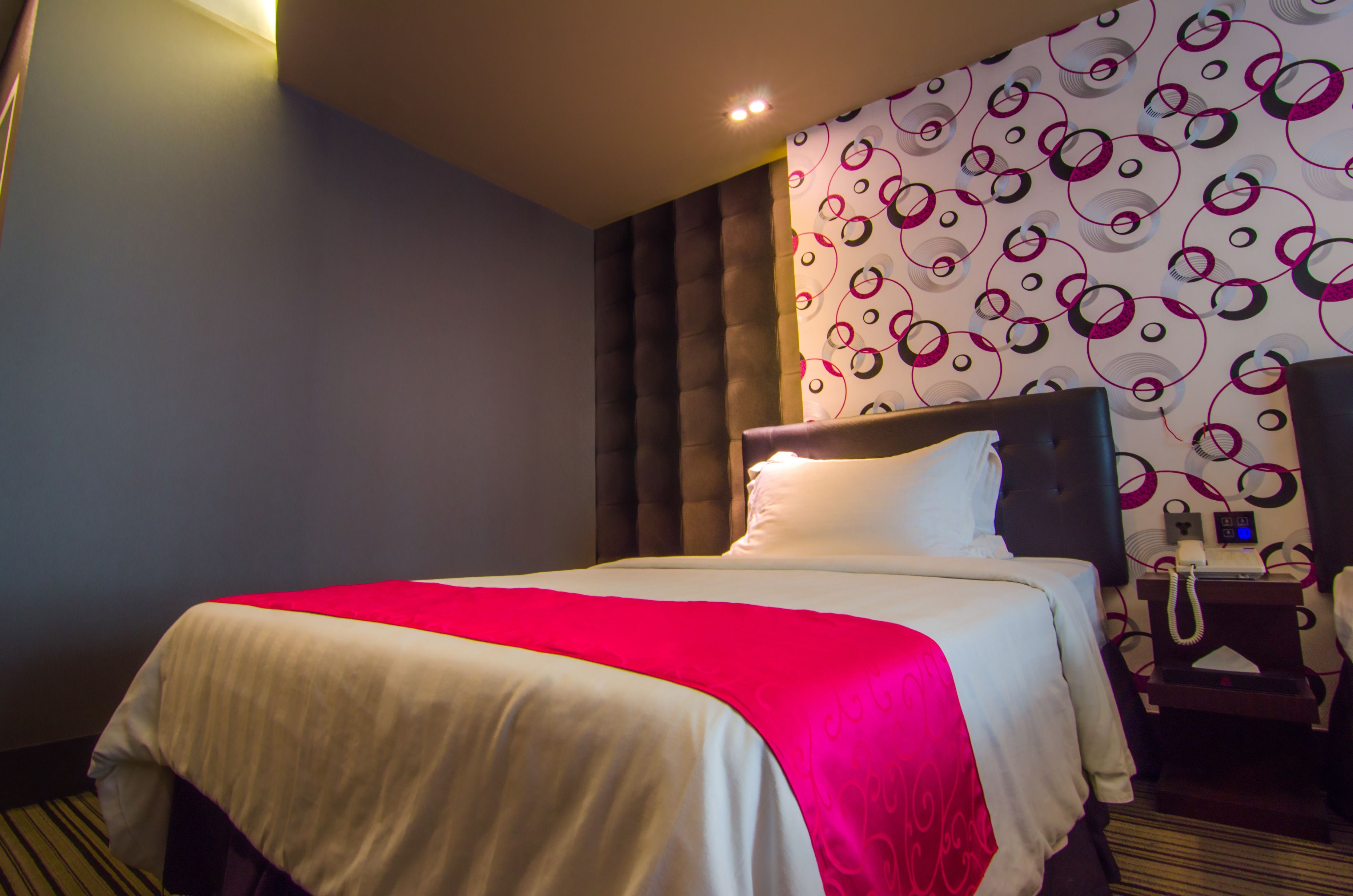 A hotel room with a bed, a headboard, a bedside table with a phone, and a patterned wall.