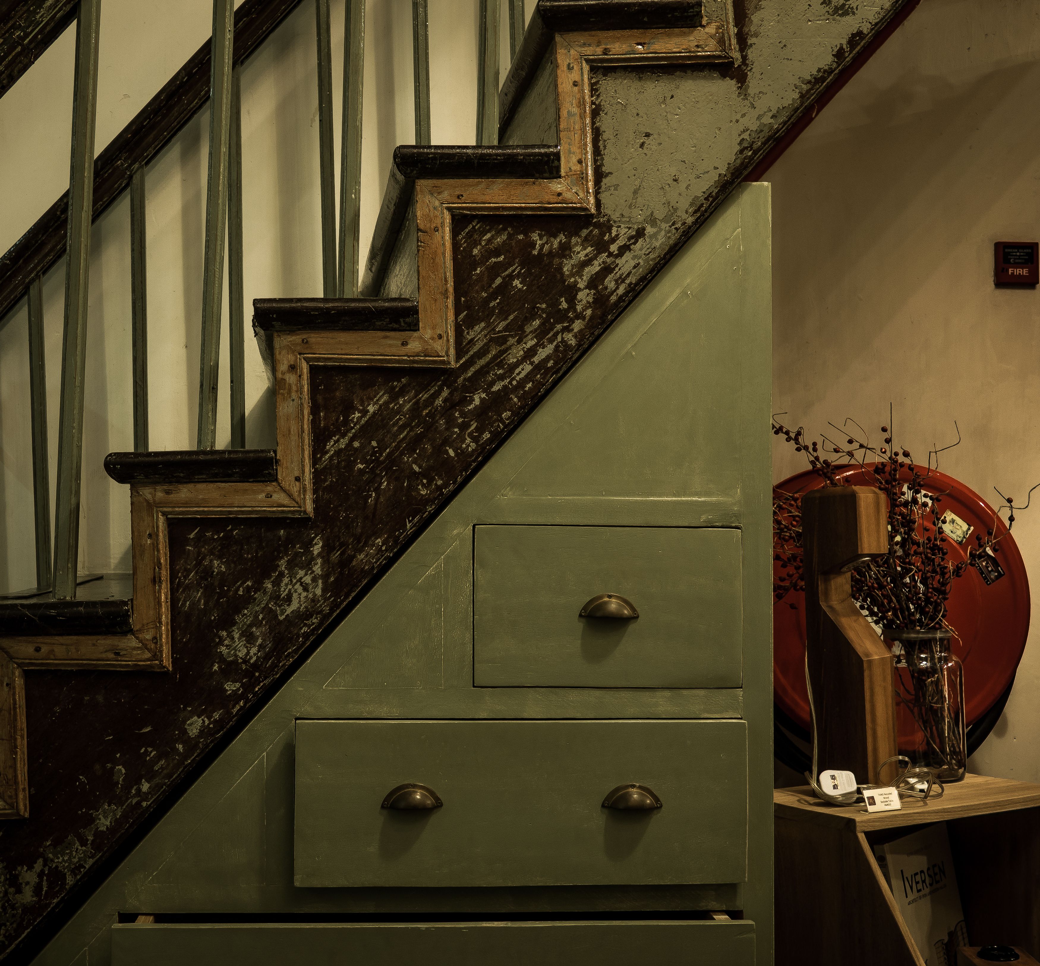 Un escalier avec un meuble vert à sa base, un objet circulaire rouge sur la droite et une petite enseigne avec du texte.