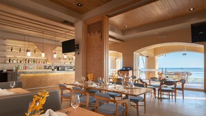 Un restaurante con zona de bar, mesas y sillas para comer. Se ve el océano a través de amplios ventanales.