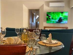 Una mesa de comedor preparada con vasos, platos, cubiertos, una vela y una servilleta decorativa. En el fondo se ve un televisor montado en la pared.