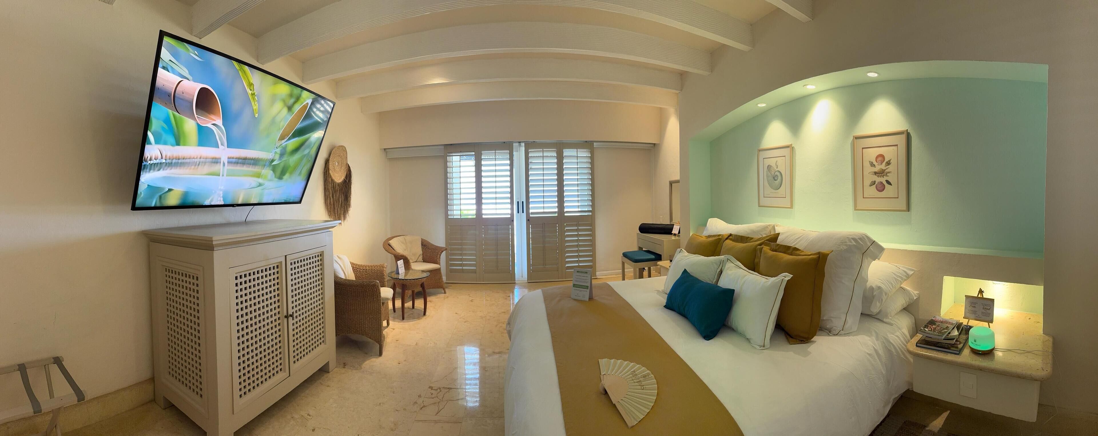 A hotel room with a flat-screen TV mounted on the wall, a bed with multiple pillows, a bedside table with a lamp, and a view of the outside through the window.