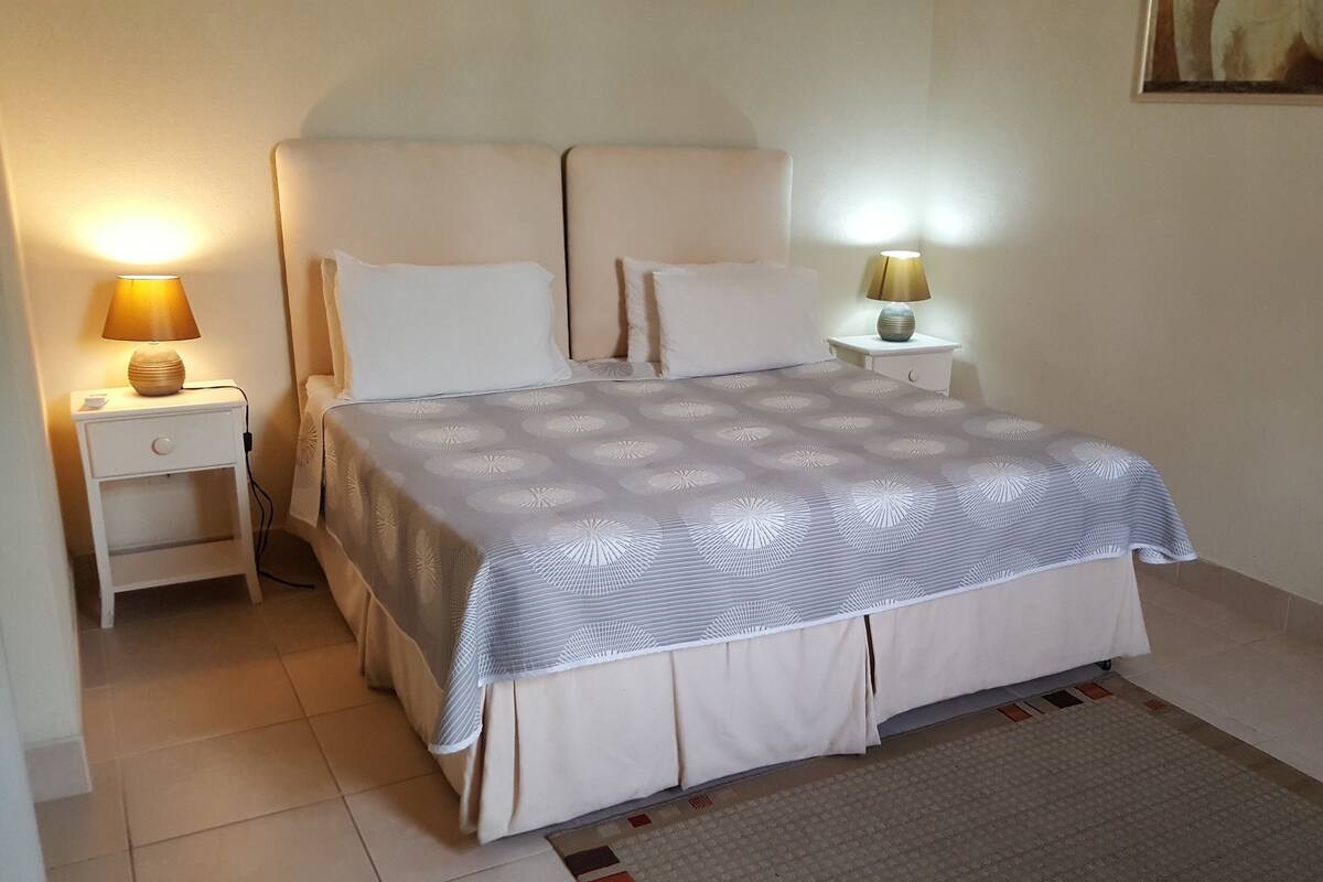 Una cama bien hecha con una colcha estampada, dos lámparas de noche y una mesita de noche con lámpara.