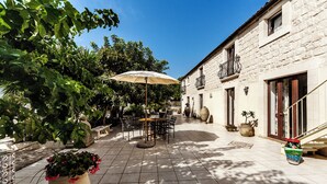 Une terrasse extérieure avec une table et des chaises sous un grand parasol, entourée de plantes en pot et d’un bâtiment en pierre.