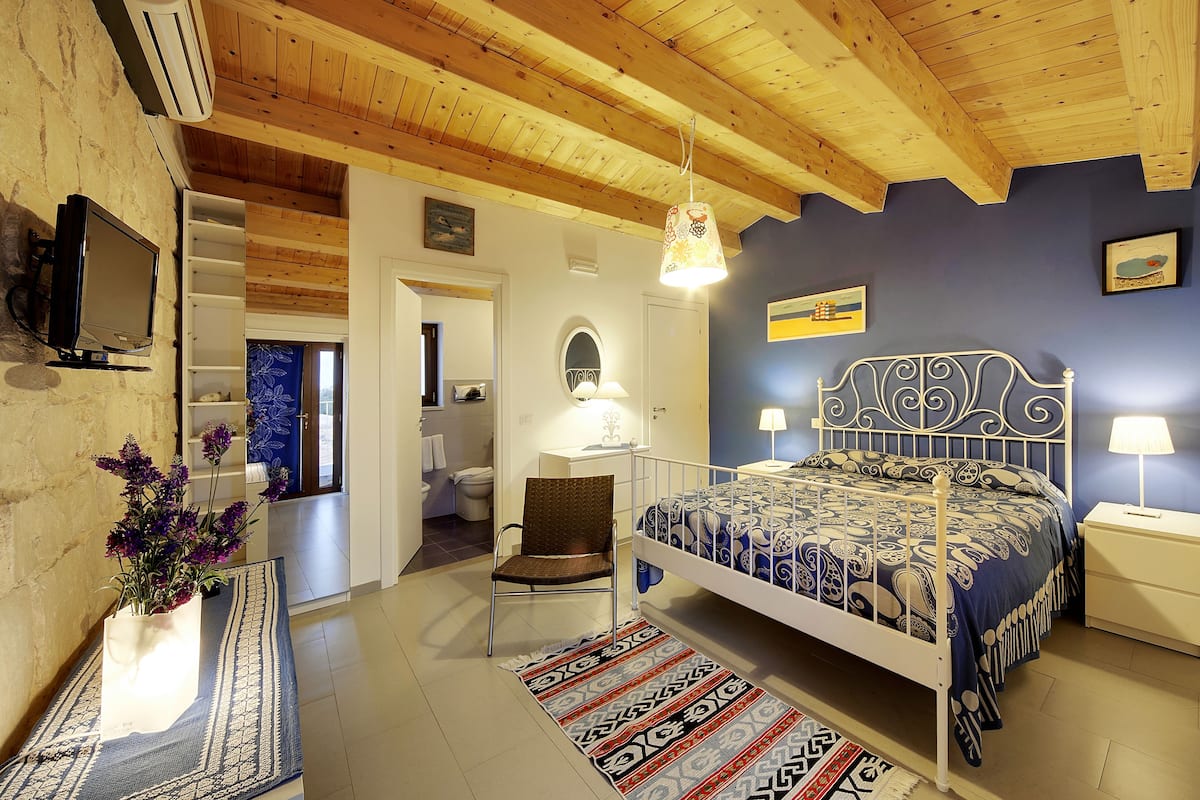 Ein Schlafzimmer mit Holzboden, einem Bett mit gemusterten Textilien, einem Nachttisch, einem Sessel, einem Schreibtisch, einem Fernseher und einem durchgehenden Türrahmen sichtbaren Badezimmer.