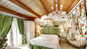 Une chambre avec un plafond en bois, une tête de lit recouverte d’un tissu vert, un fauteuil à motifs floraux et un lustre.