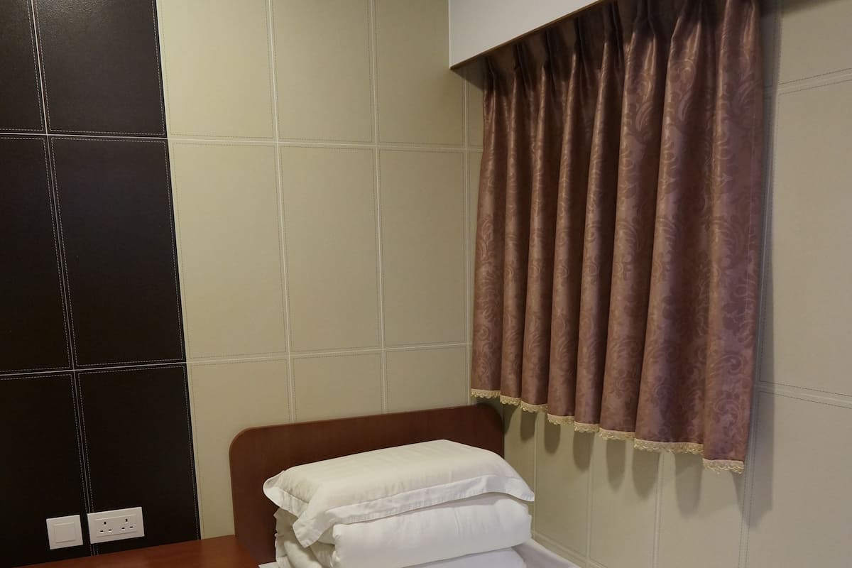 A small, single-bed room with a wooden headboard, a bedside table with a lamp, and a window with patterned curtains.