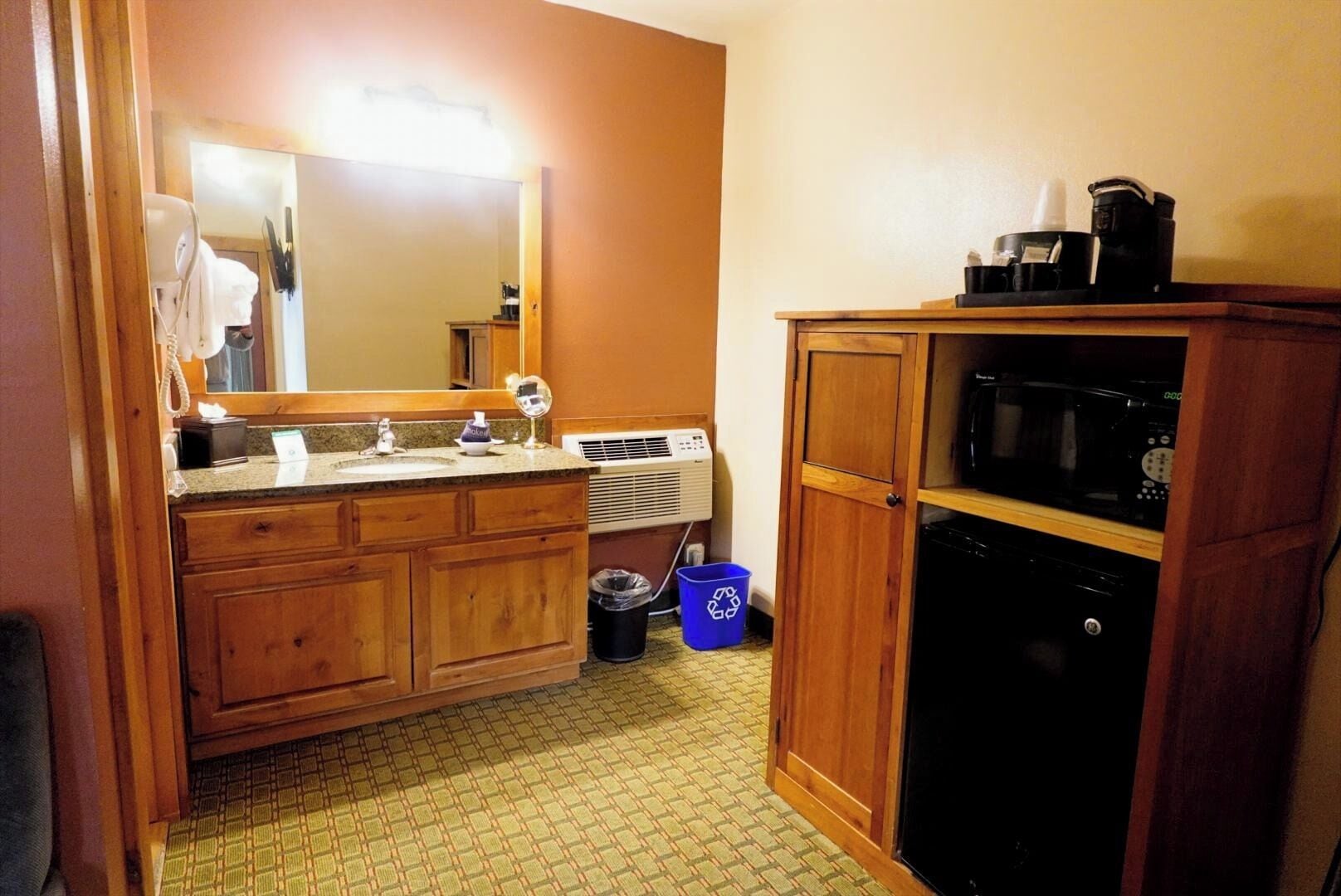 A bathroom with wooden cabinets, a sink, a mirror, a trash bin, and a microwave.