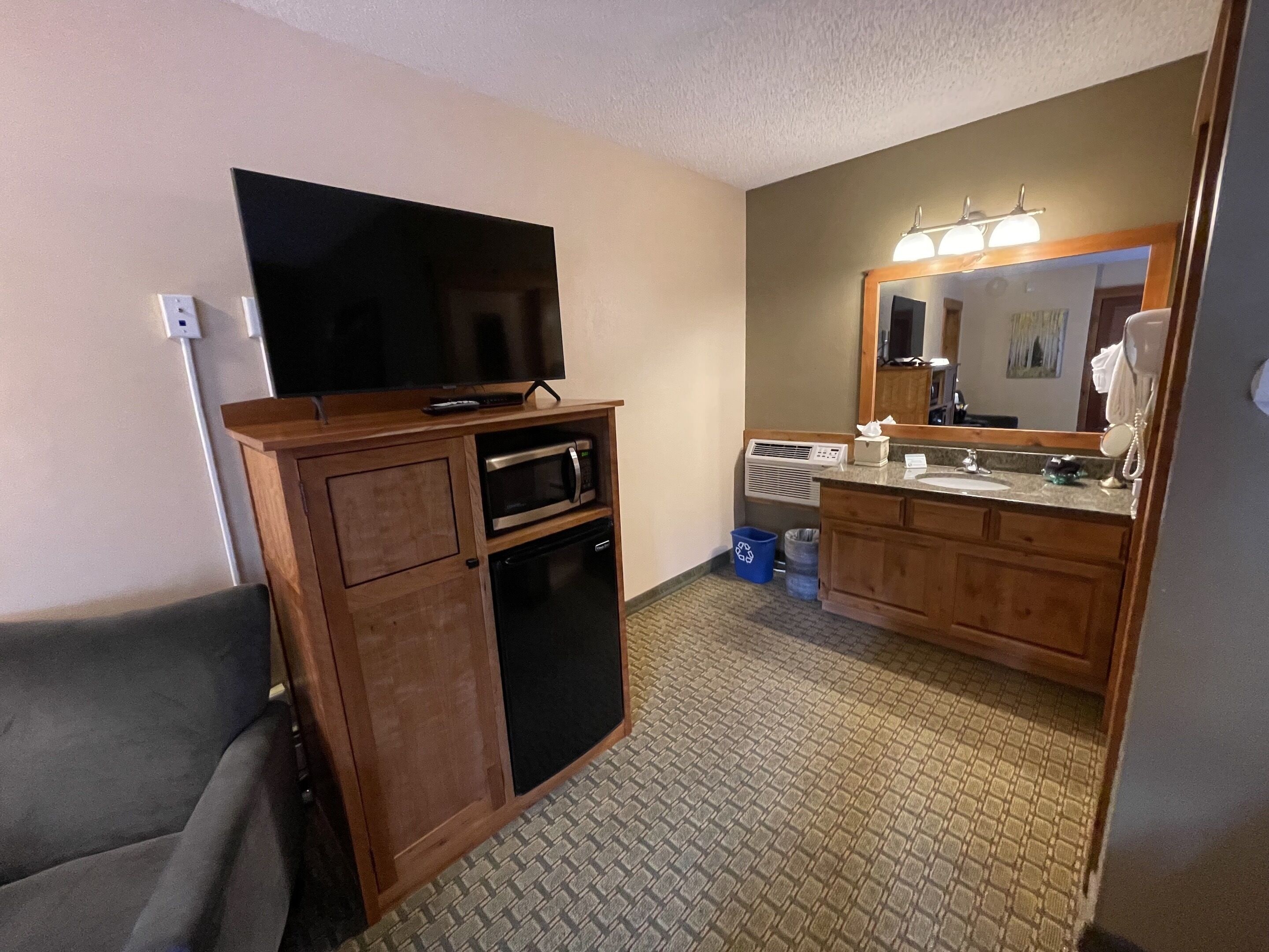 Habitación de hotel compacta con kitchenette, televisión de pantalla plana, baño con lavamanos y espejo, y alfombra estampada.