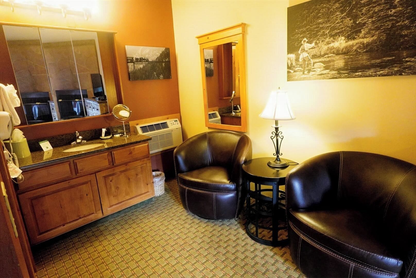 A hotel room with a wooden vanity, a large mirror, a small round table with a lamp, two leather armchairs, and a painting on the wall.