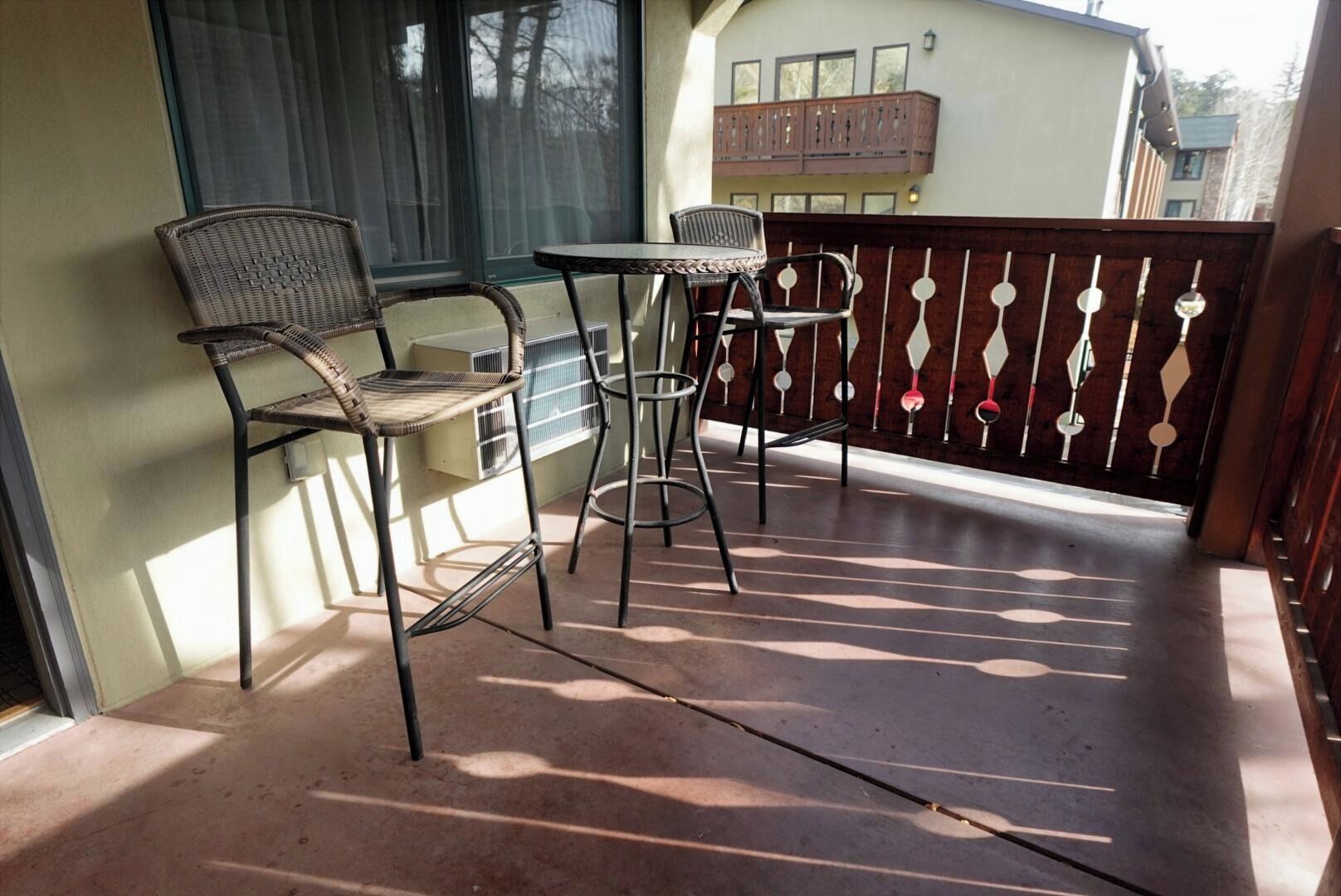 Een balkon met twee rieten stoelen en een klein tafeltje, een houten hek en een gebouw op de achtergrond.