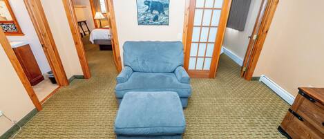 Une pièce avec un fauteuil bleu et un pouf, une armoire en bois et un tableau encadré au mur.
