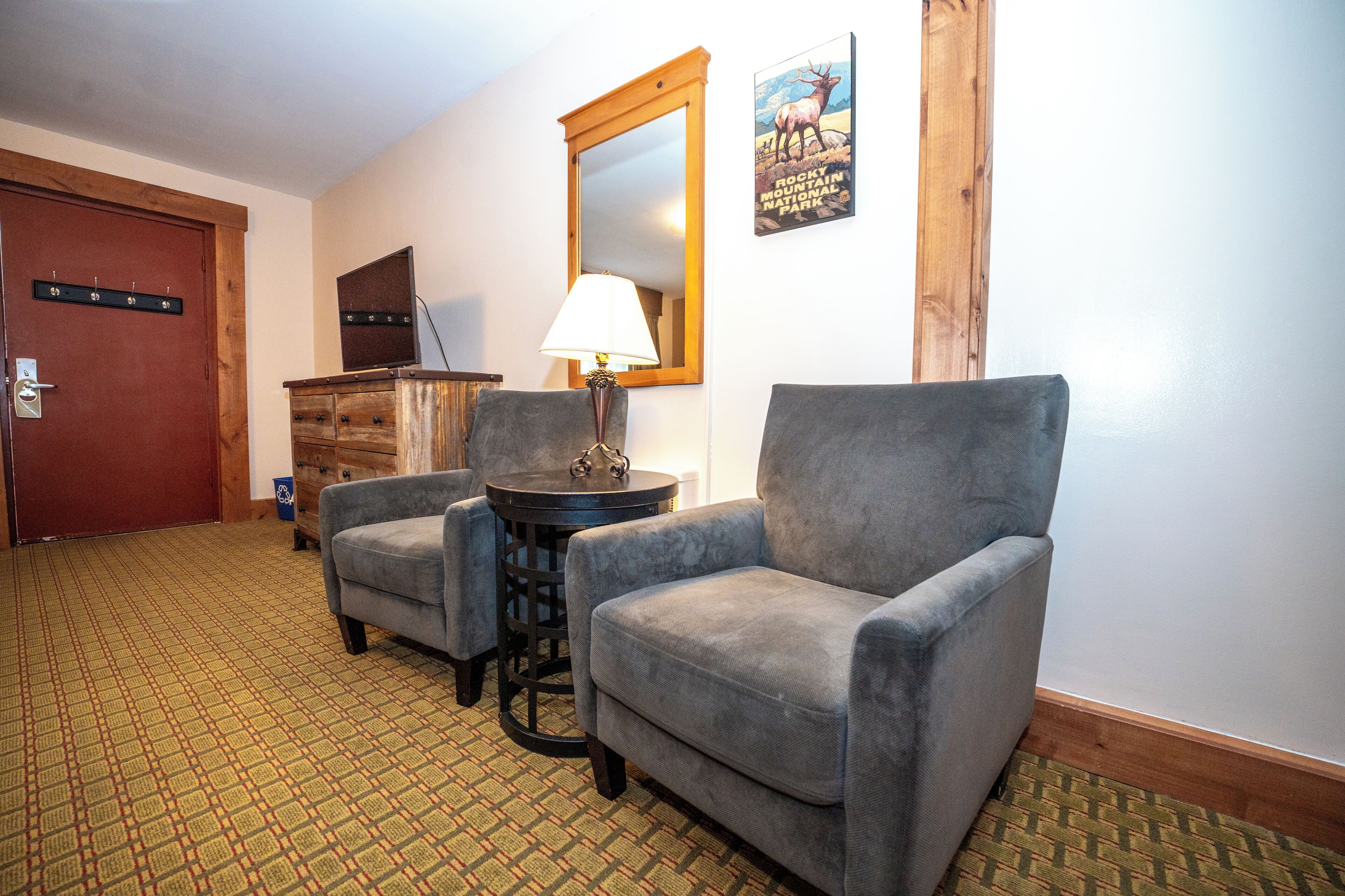 A hotel room with a wooden door, a mirror, a lamp, two armchairs, a small table, and a patterned carpet.