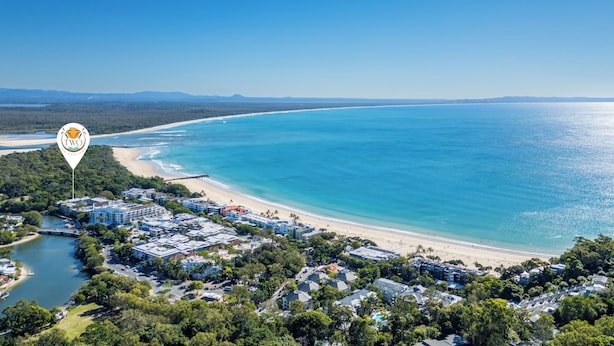 An aerial view of a coastal area with a beach, clear blue water, and a mix of residential and commercial buildings. - 2 Hastings Street (Noosa Heads)