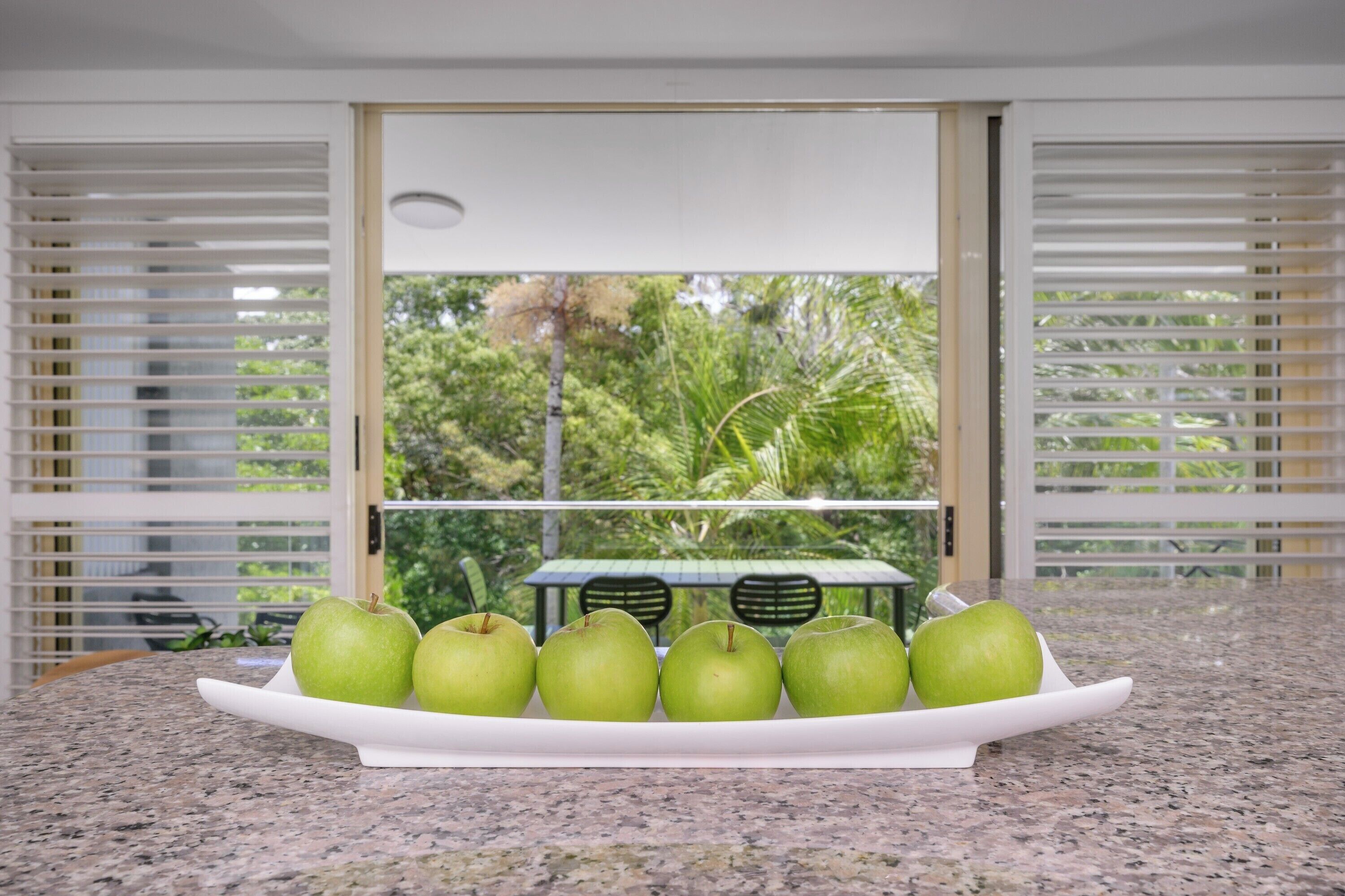 A kitchen counter with a white plate holding six green apples, a window with blinds, and a view of greenery outside.