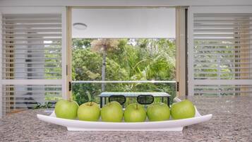 A kitchen counter with a white plate holding six green apples, a window with blinds, and a view of greenery outside.
