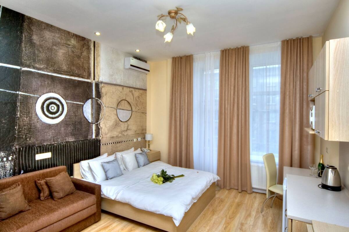 A modern hotel room with a large bed, a sofa, a desk with a kettle, and a window with curtains.