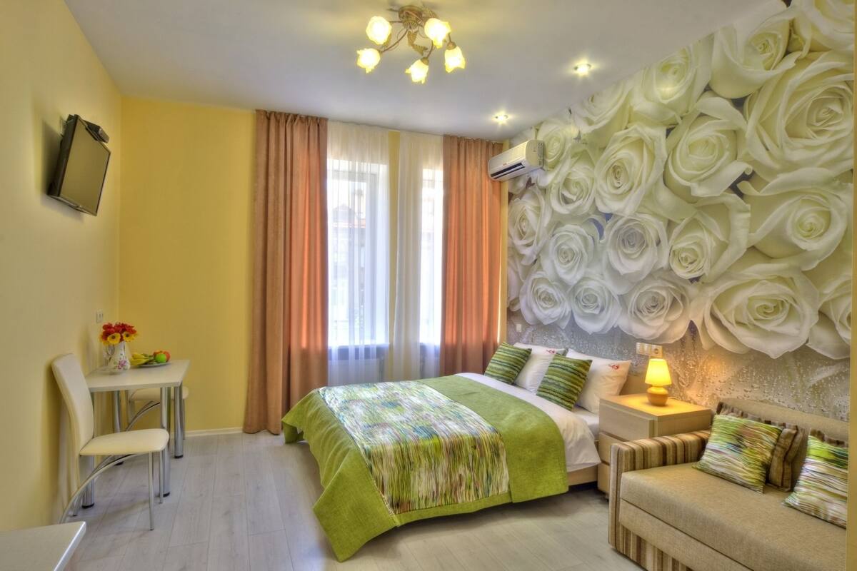 A hotel room with a bed, a chair, a table with flowers, a wall with a floral design, and a ceiling light.