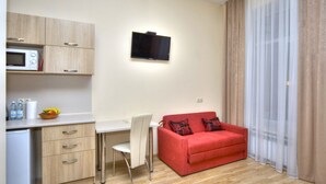 A compact living space with a kitchenette, a flat-screen TV mounted on the wall, a red sofa with patterned cushions, and a large window with sheer curtains. - Fire Inn (Kyiv)