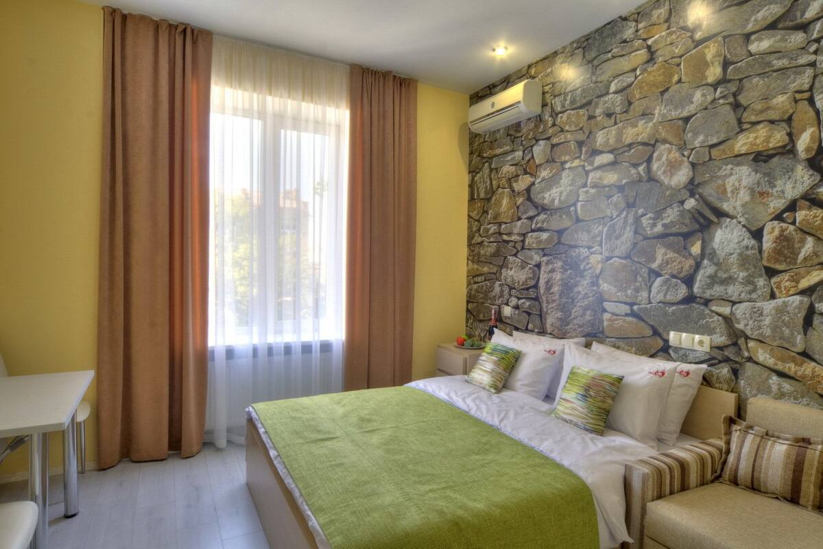 A bedroom with a stone wall, a bed with pillows, a table, and a window with curtains.