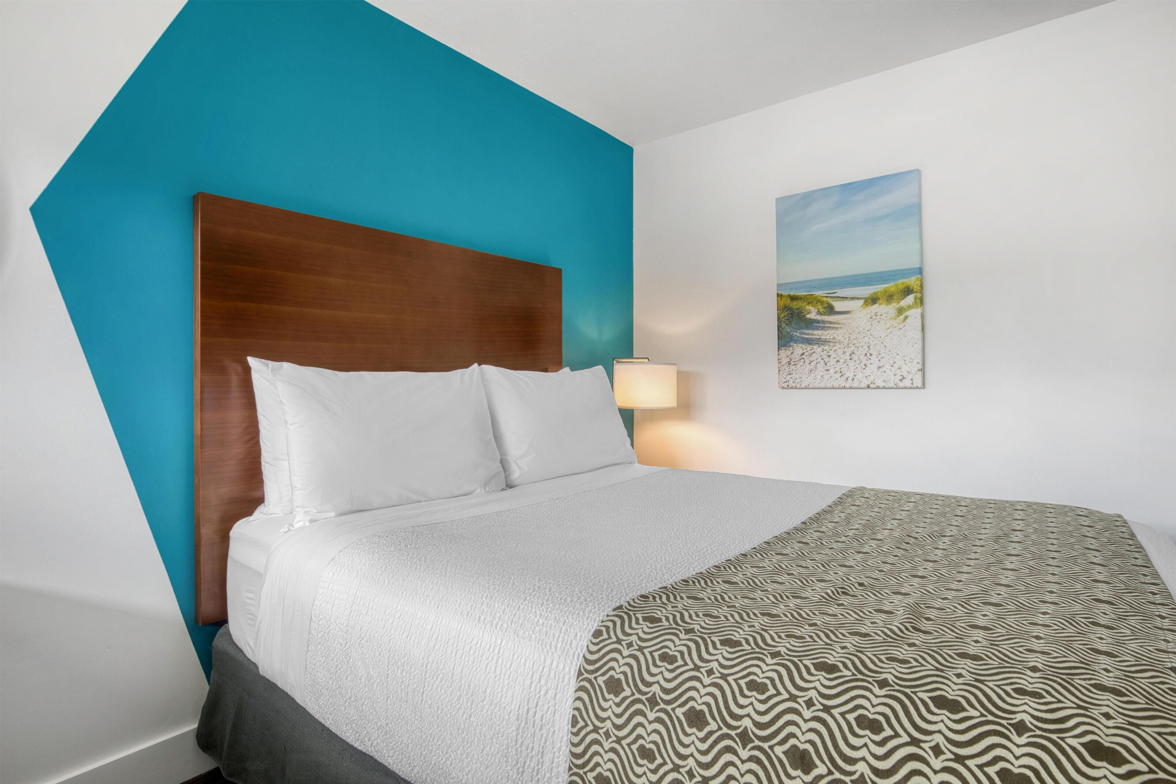 Habitación de hotel con cama, mesita de noche, lámpara y un cuadro con una escena de playa en la pared.