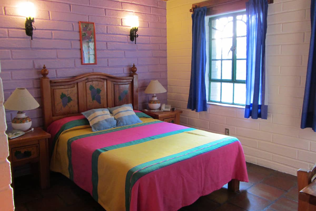 Una cama con una colcha colorida y dos almohadas, una mesita de noche con una lámpara, una luz empotrada en la pared y una ventana con cortinas.