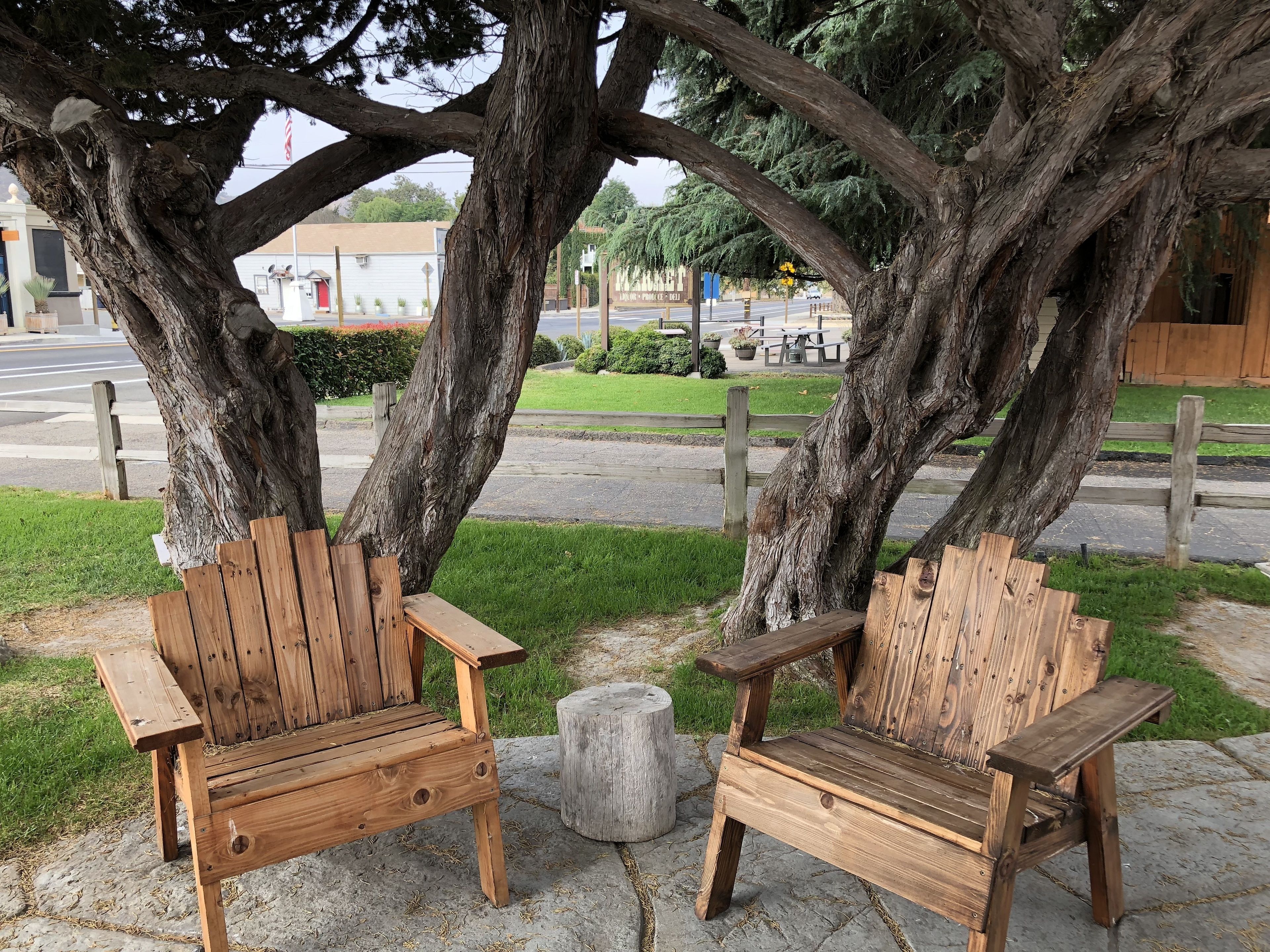 Two wooden Adirondack chairs under large trees on a paved area with a grassy lawn and a road in the background.