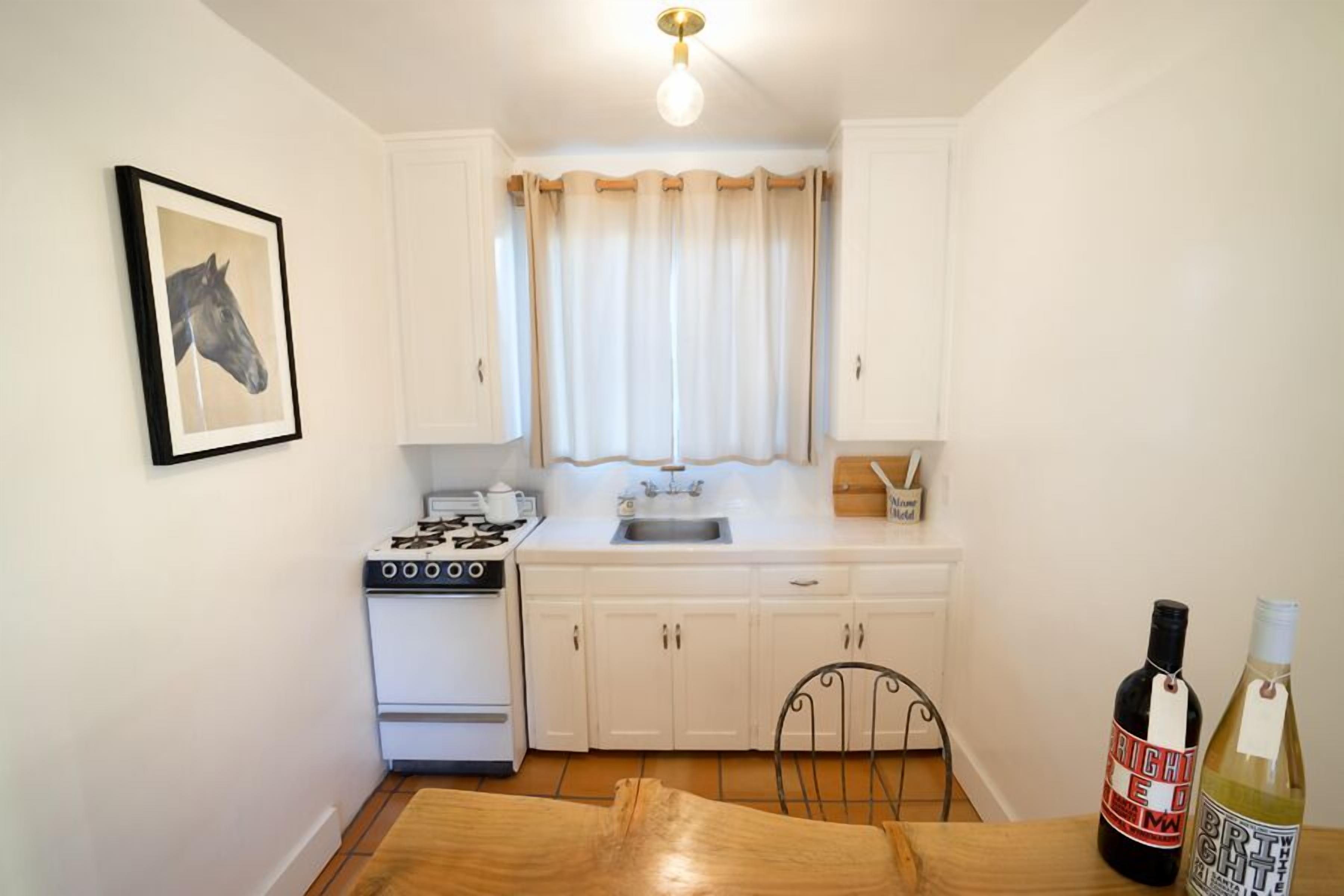 Une petite cuisine avec des meubles blancs, une cuisinière, un évier et une table à manger. Il y a deux bouteilles de vin sur la table.