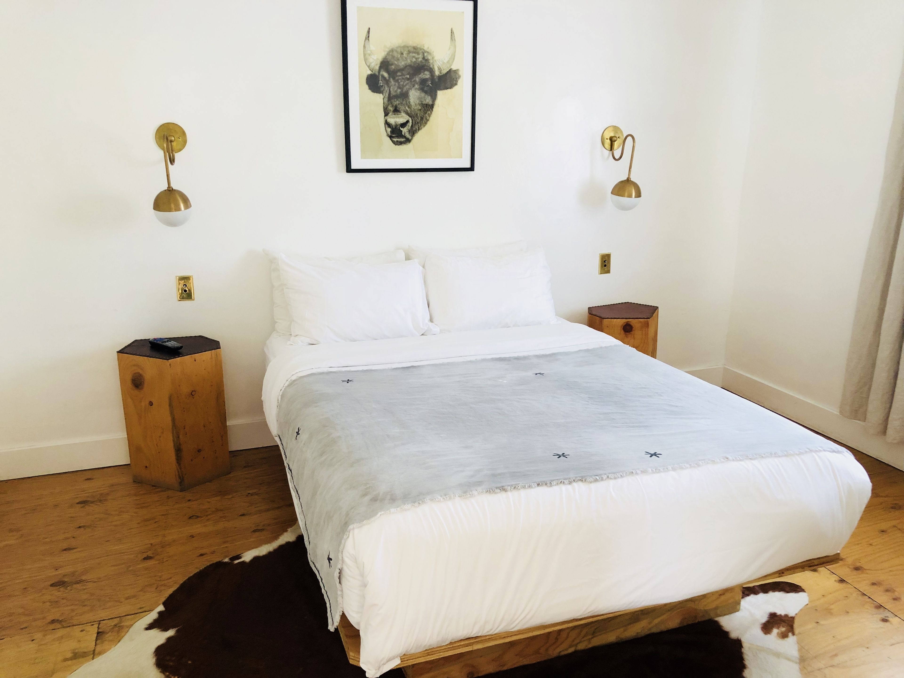 A bedroom with a bed, white pillows, a grey blanket, wooden nightstands, a cowhide rug, and a bull-themed artwork on the wall.