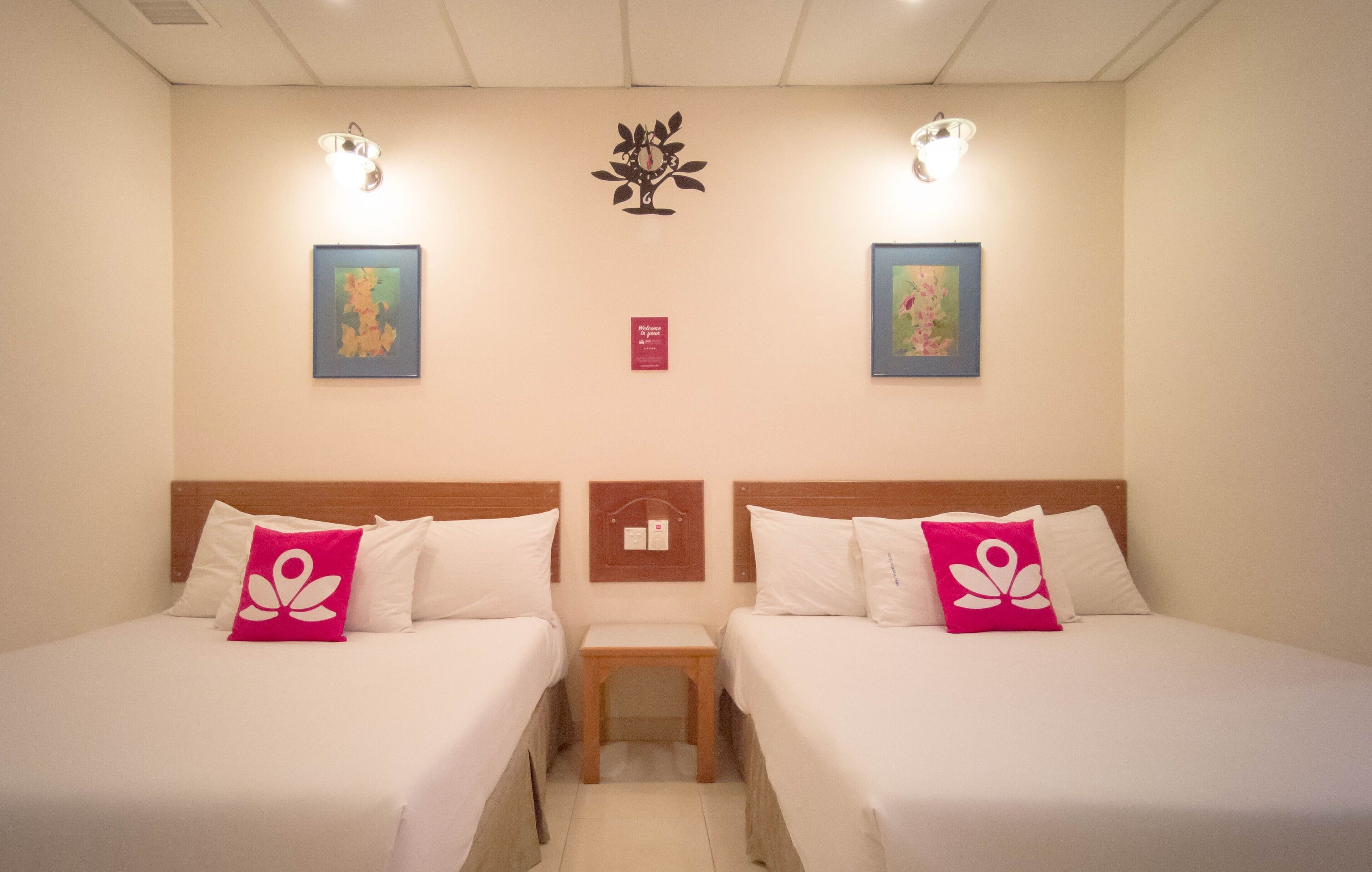 A hotel room with two beds, each with white bedding and a pink pillow. There are wall-mounted light fixtures and framed artwork above the beds.
