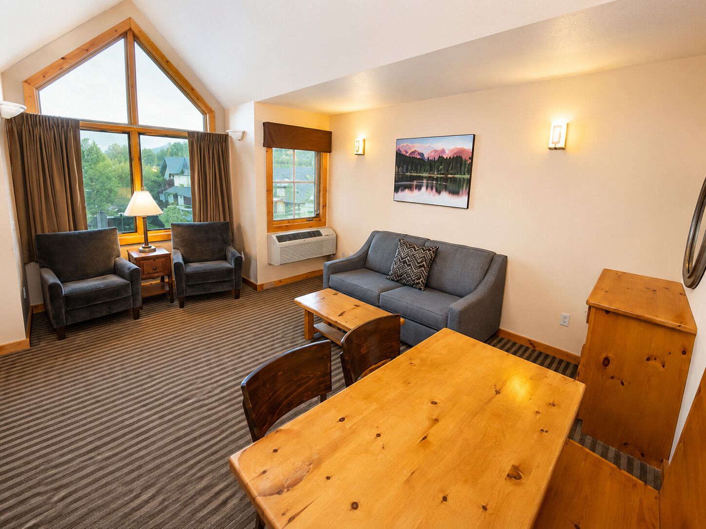 A hotel room with a large window, a sofa, two armchairs, a wooden table, and a small side table with a lamp.