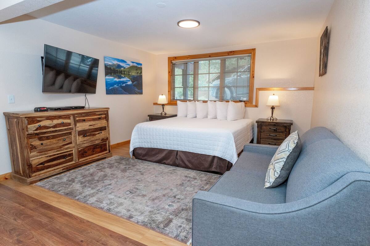 Quarto com cama, sofá cinza, cômoda de madeira e duas luminárias de parede.