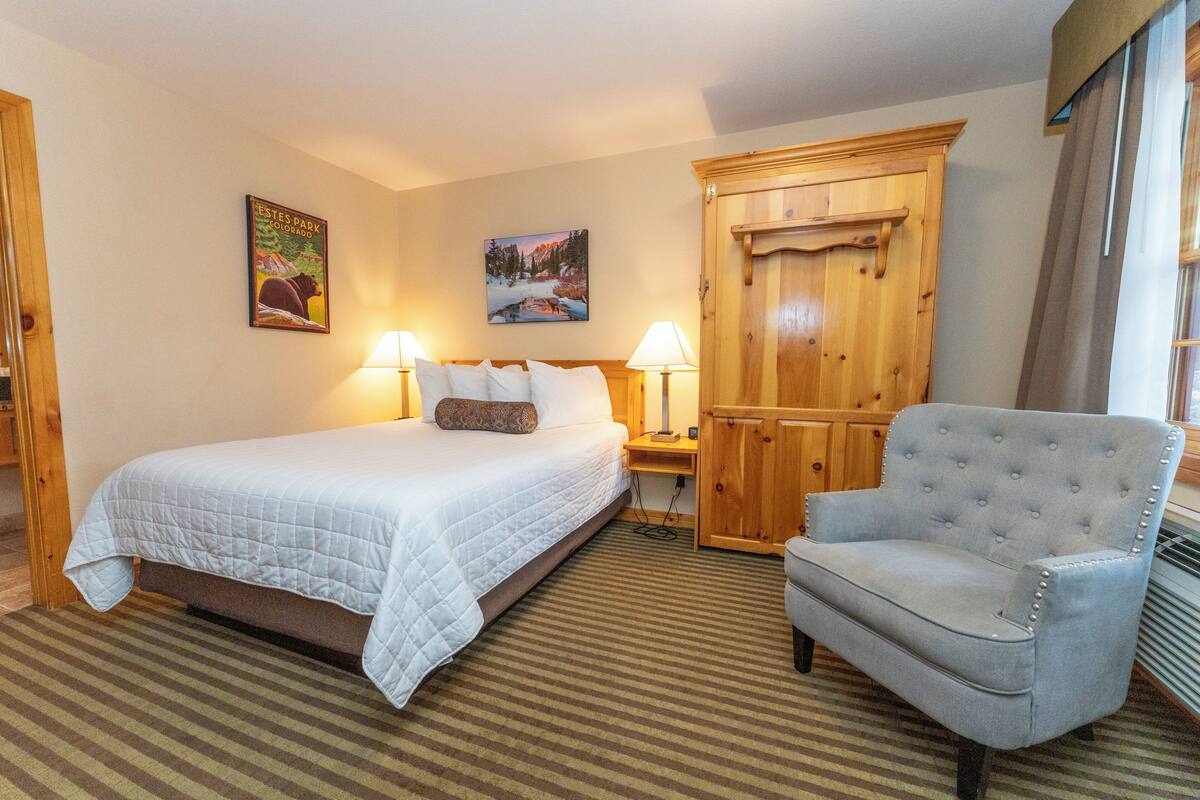 Quarto de hotel com uma cama, duas luminárias de cabeceira, um guarda-roupa de madeira e uma poltrona cinza.
