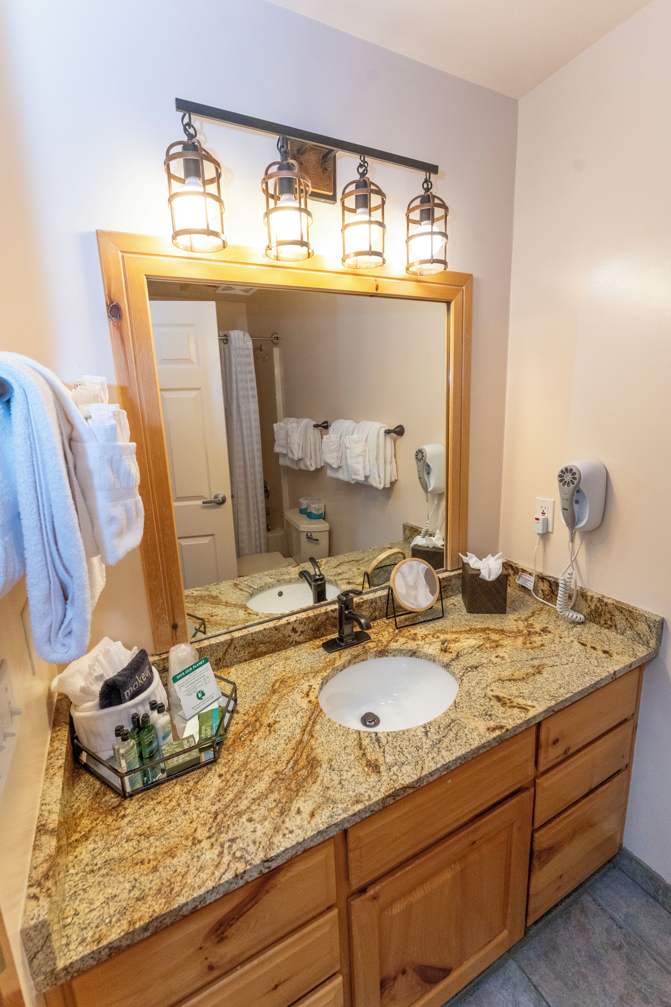 A bathroom with a granite countertop, wooden cabinets, a large mirror, a hairdryer, and a towel rack.