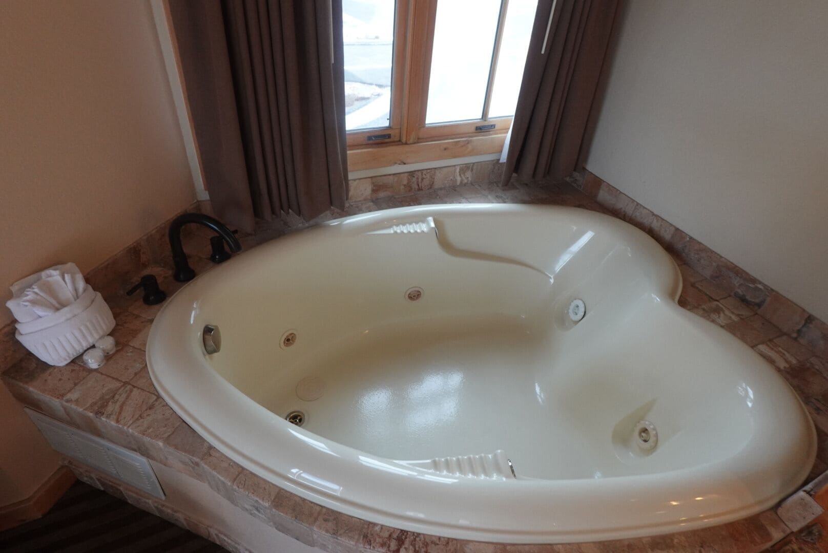 A large, oval-shaped bathtub with a faucet and a drain, situated in a bathroom with a stone-tiled floor and a window with curtains.