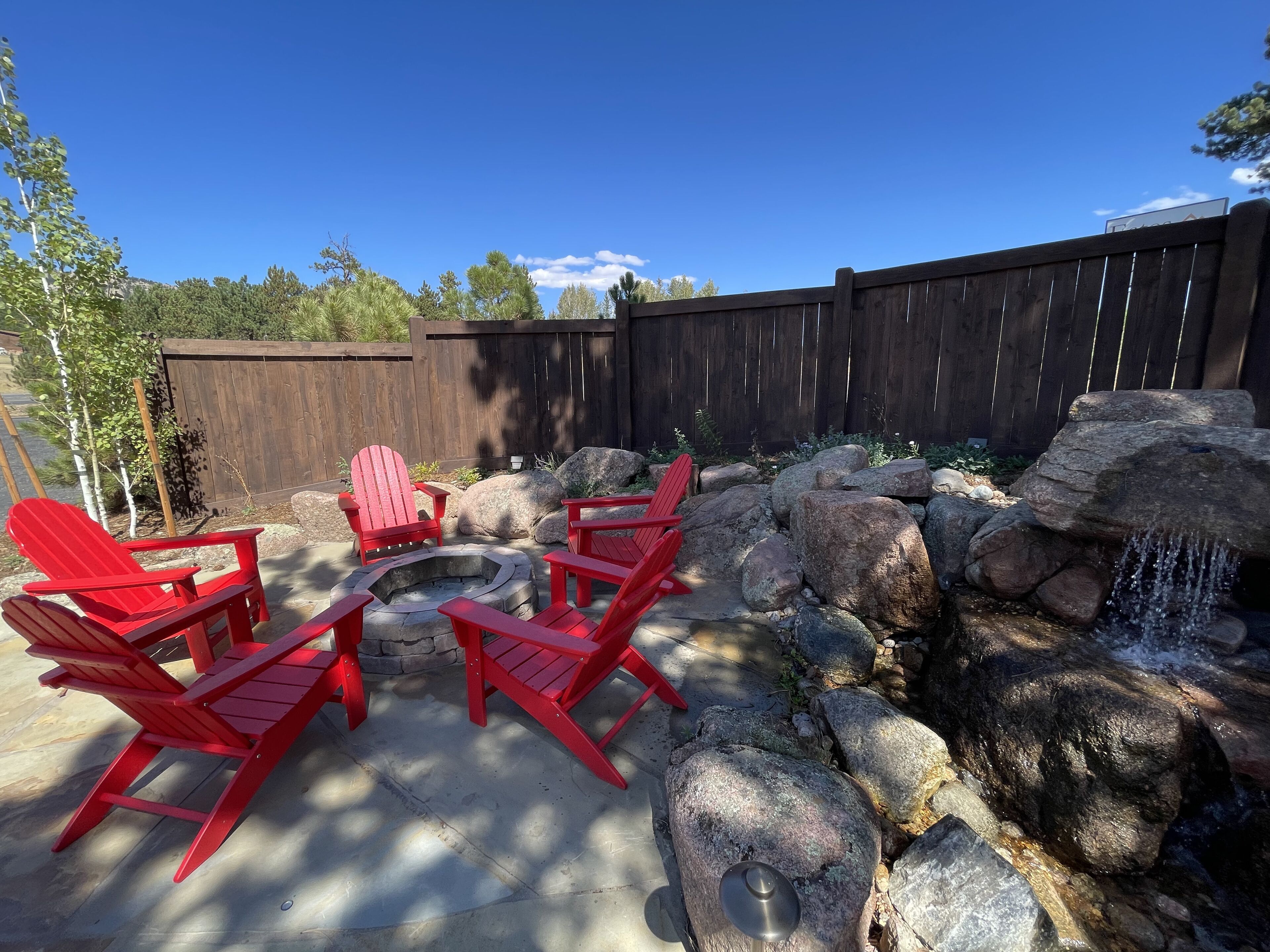 A backyard with a fire pit, red Adirondack chairs, and a rock waterfall.