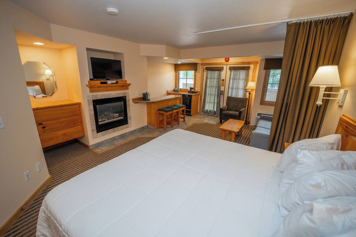 Quarto de hotel com uma cama grande, lareira, televisão, pequena cozinha e área de estar com uma cadeira e mesa.